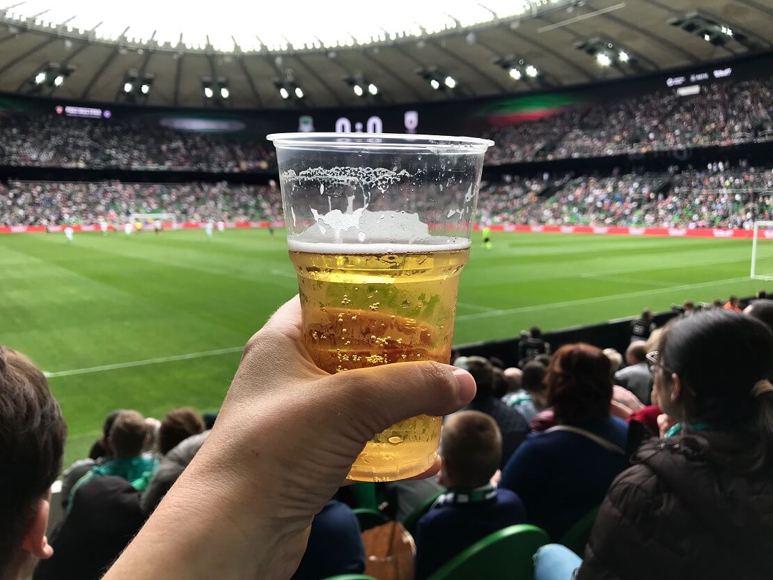 Bierbecher im Stadion Hand mit einem Plastikbecher mit Bier, dahinter Zuschauer im Fußballstadion